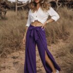 Kobieta w białej koszuli i fioletowych spodniach z wysokim stanem i rozcięciami, pozująca na wydmach.
