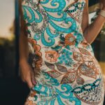 Bliskie ujęcie na wzorzysty materiał letniej sukienki z motywem paisley, turkusowymi i pomarańczowymi kwiatami oraz złotym łańcuszkiem