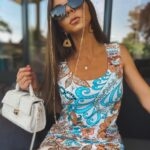 Kobieta w kolorowej letniej sukience w wzór paisley, w okularach przeciwsłonecznych na złotym łańcuszku, trzymająca małą białą torebkę