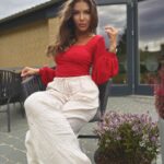 Kobieta w czerwonym body i szerokich lnianych spodniach siedzi elegancko w ogrodzie.