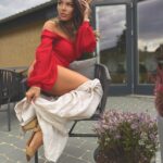 Kobieta w czerwonym body z opadającym ramieniem patrzy w bok, z wiatrem we włosach.