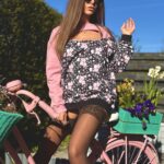 Modelka Sensual Woman w różowo-czarnej bluzie z kwiatami i wycięciem siedząca na różowym rowerze z koszykami kwiatów